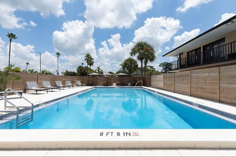 Resort swimming pool with depth markings, lounge chairs, palm trees, and wooden privacy fence