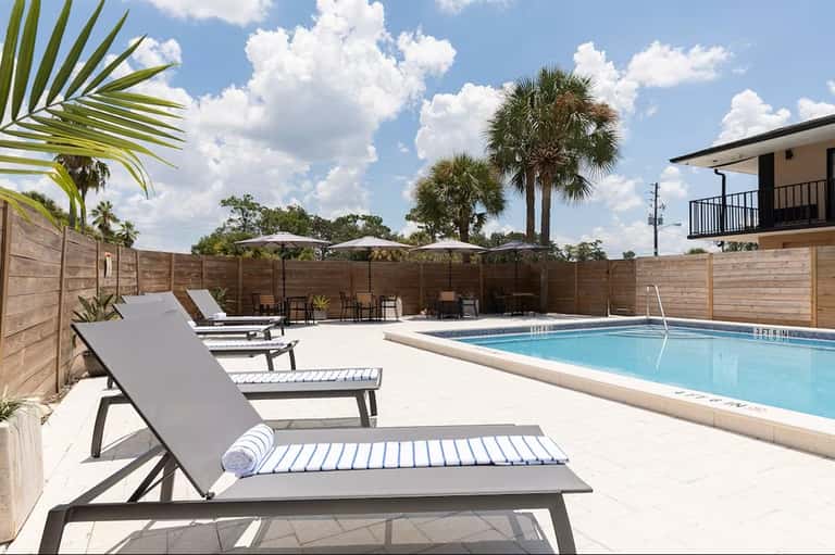 Modern pool deck with white lounge chairs, wooden privacy fence, palm trees, and clear blue pool