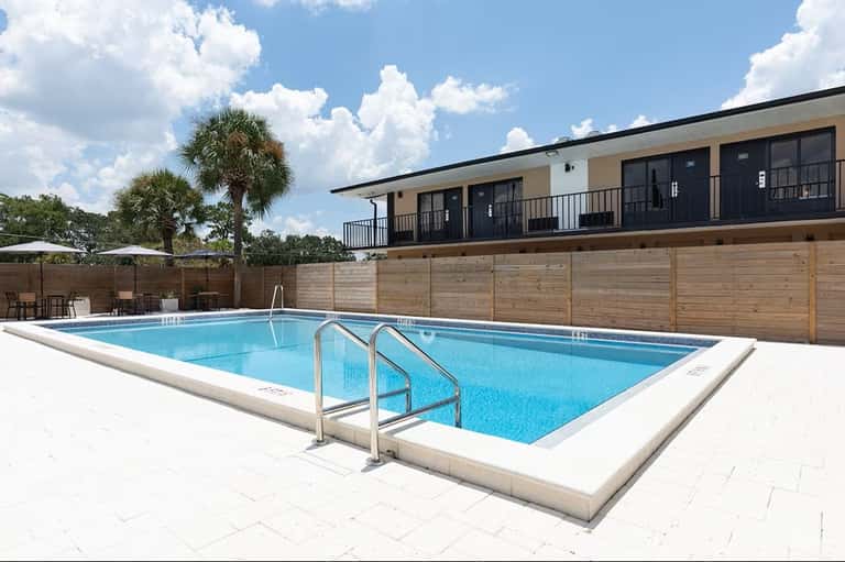 Rectangular swimming pool with white deck, metal railings, palm trees, wooden fence, and two-story building with balconies
