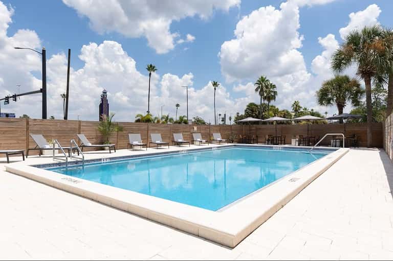 Rectangular resort pool with white lounge chairs, wooden privacy fence, and palm trees against blue sky