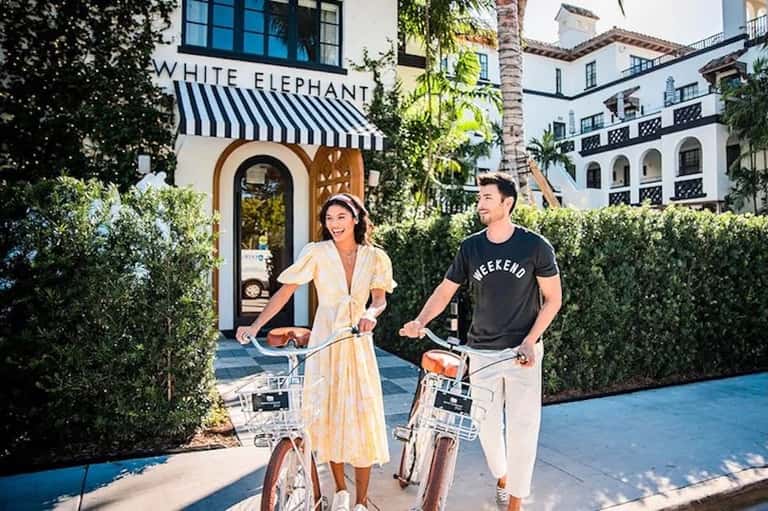 Couple riding bicycles past White Elephant storefront in resort courtyard with Mediterranean architecture