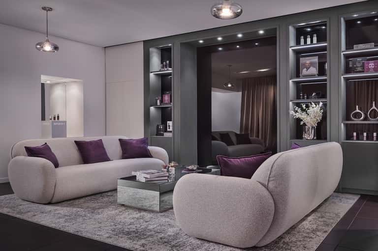 Luxury spa lounge with curved beige seating, dark shelving with beauty products, and soft pendant lighting