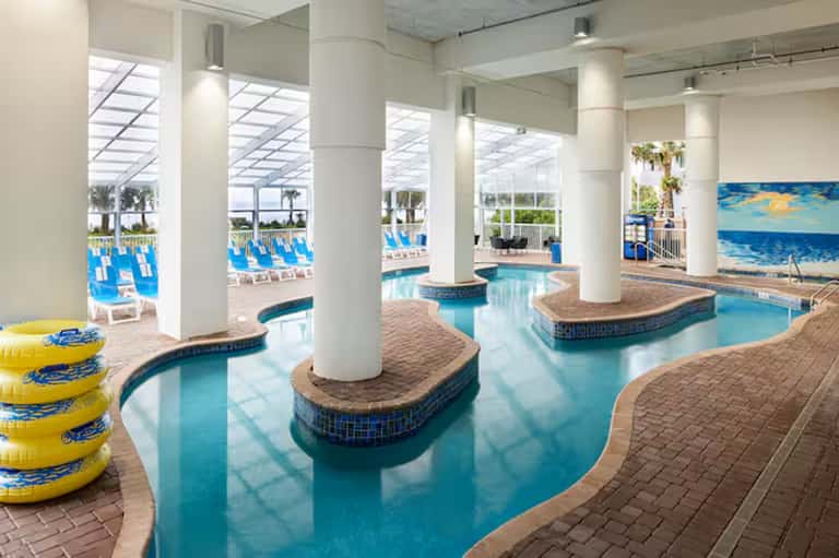 Indoor resort pool with bright skylights, multiple pools, loungers, and white support columns
