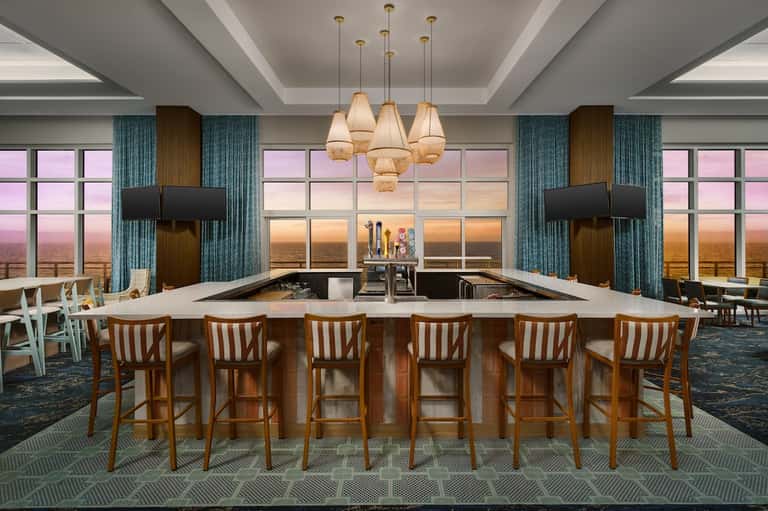 Modern rooftop bar with expansive windows, fireplace, pendant lights, and sunset ocean view