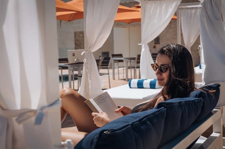 Woman reading under white cabana curtains on lounger by poolside