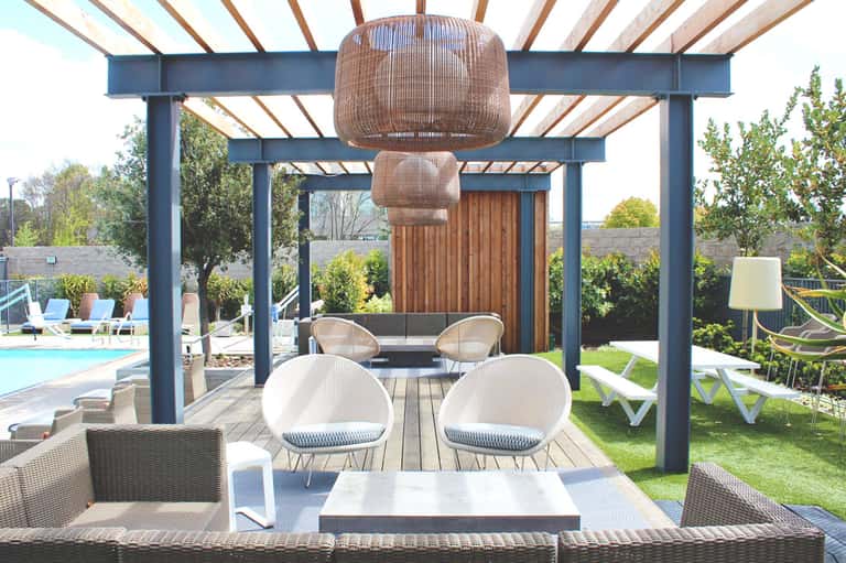 Modern pergola patio with woven pendant lights, white chairs around fire table, pool and greenery visible.