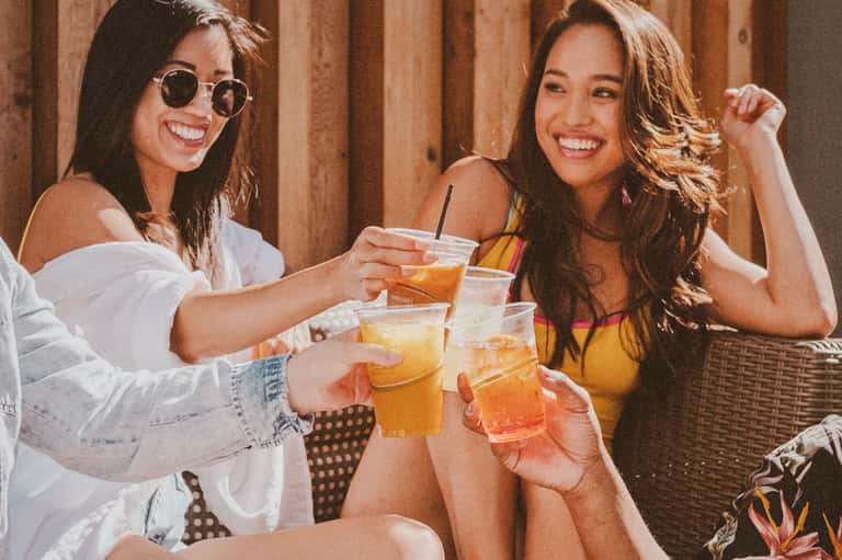 Two women toasting with colorful drinks at an outdoor venue with warm lighting and tropical decor