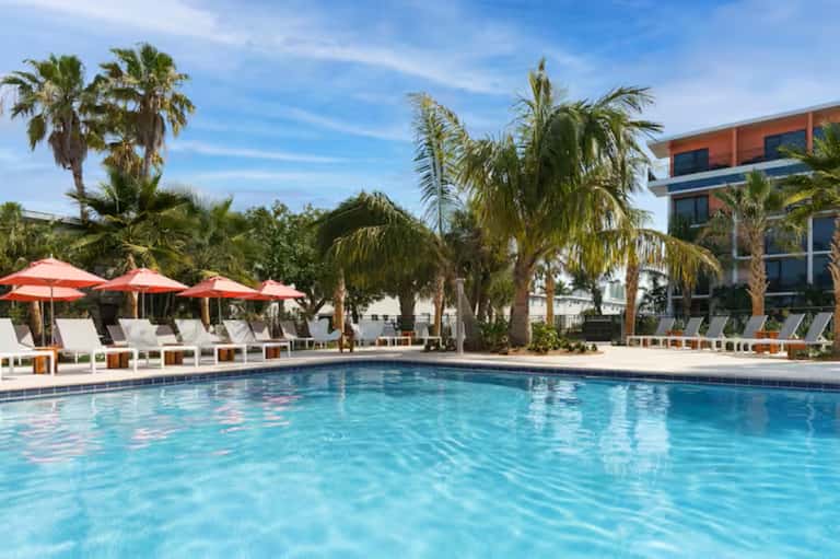 Resort pool surrounded by palm trees, red umbrellas, lounge chairs, and modern building