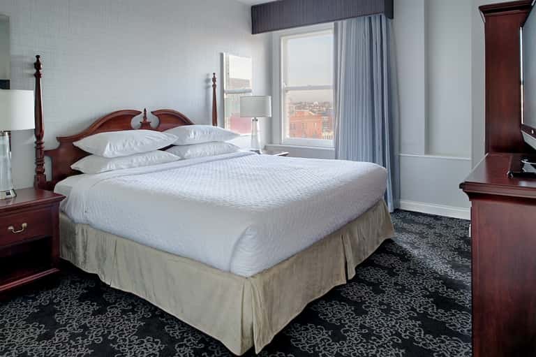Upscale hotel bedroom with queen bed, burgundy headboard, floor-to-ceiling windows, and city view