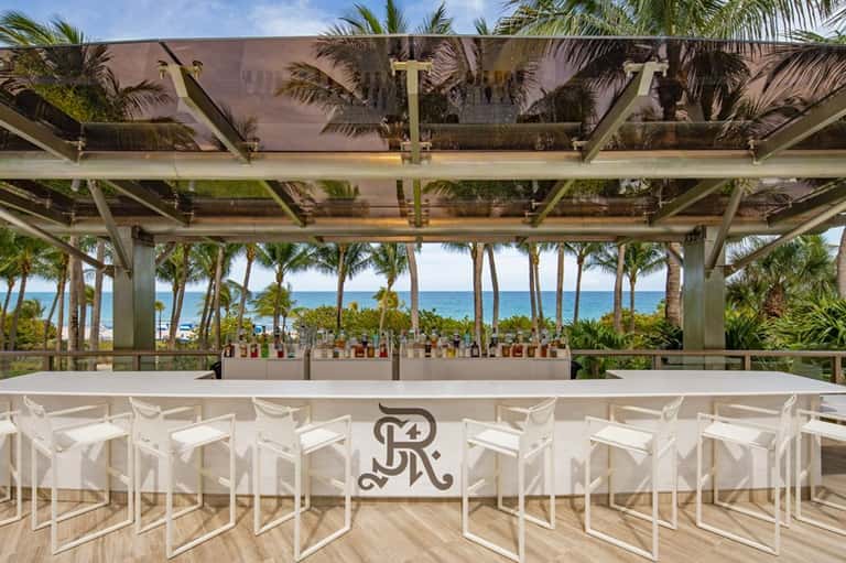 Beachfront bar with white chairs, periscope ceiling, palm trees, and ocean view in background