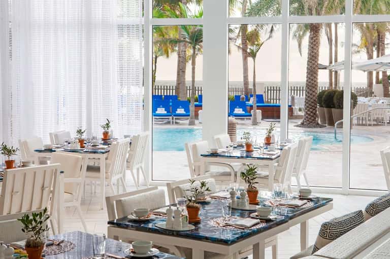 Upscale dining room with white chairs, ocean-view windows, and resort pool visible outside