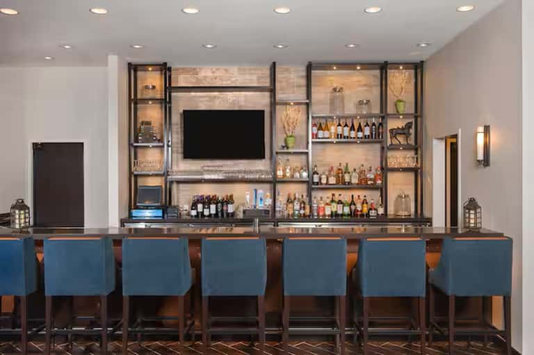 Modern hotel bar with illuminated shelving displaying bottles, TV screen, and blue upholstered seating