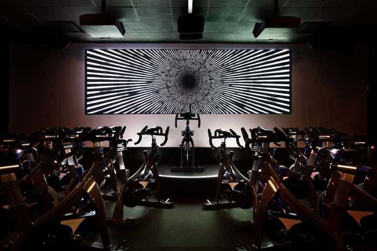Indoor spin studio with rows of exercise bikes facing large illuminated geometric wall display screen