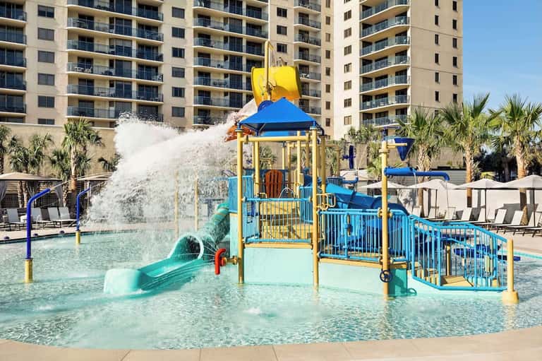 Waterpark with colorful splash pad structure, water slide, shallow pool, and resort towers backdrop