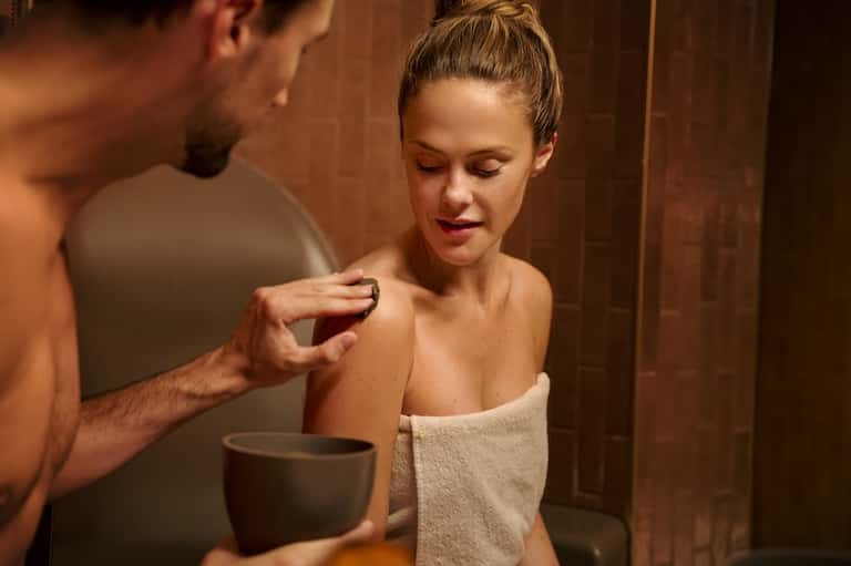 Spa treatment scene showing therapist applying product to client's shoulder in warm-toned room