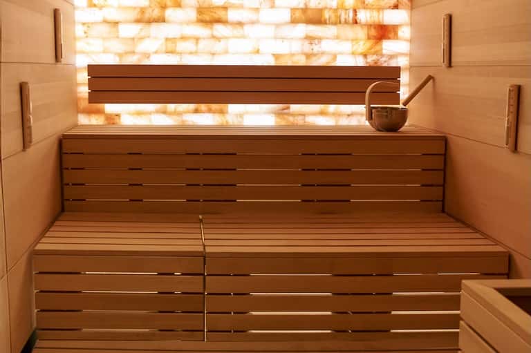 Wooden sauna interior with illuminated amber salt brick wall and traditional sauna benches and ladle