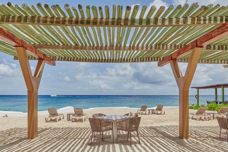 Beach lounge area with striped pergola, ocean view, lounge chairs, and dining table at sand level