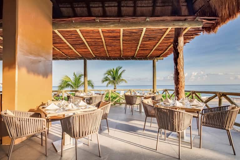 Oceanfront restaurant with woven pergola, wooden chairs, white table settings, and palm tree views