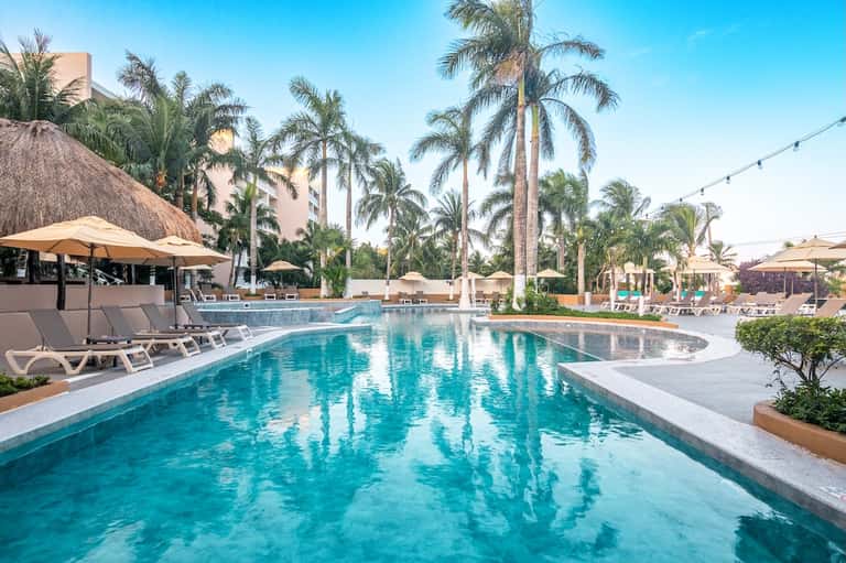 Large resort pool with palm trees, white lounge chairs, umbrellas, and high-rise building