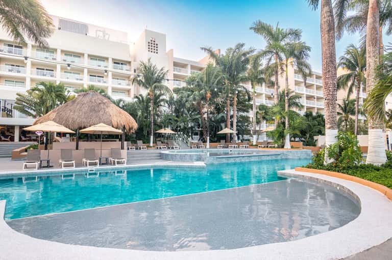 Resort pool area with thatched umbrellas, palm trees, and multi-story hotel building