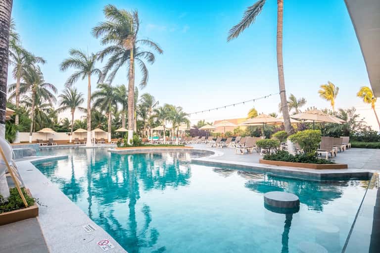 Large resort pool with turquoise water, palm trees, lounge chairs, white umbrellas, and desert landscaping