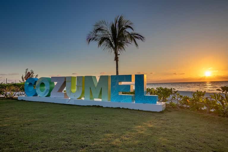 Colorful Cozumel sign on grassy beachfront at sunset with palm tree and ocean backdrop