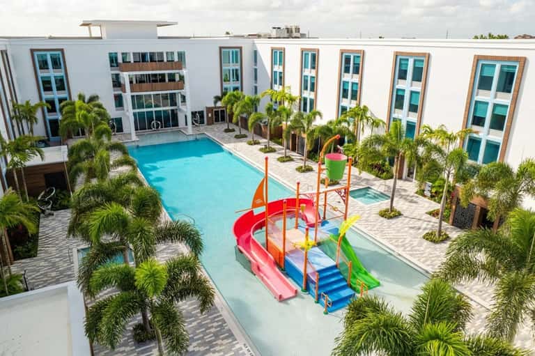 Resort courtyard with bright blue swimming pool, colorful waterslide, palm trees, white hotel buildings, and paved grounds