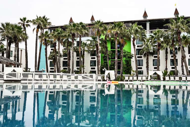 Modern resort building with white and green facade, palm trees, and reflection in calm pool water