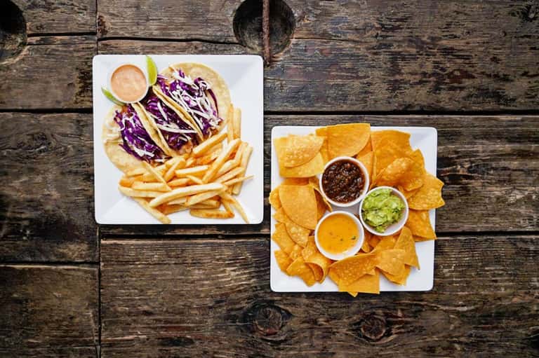 Tacos with purple cabbage and fries, and tortilla chips with salsa and guacamole on wooden table