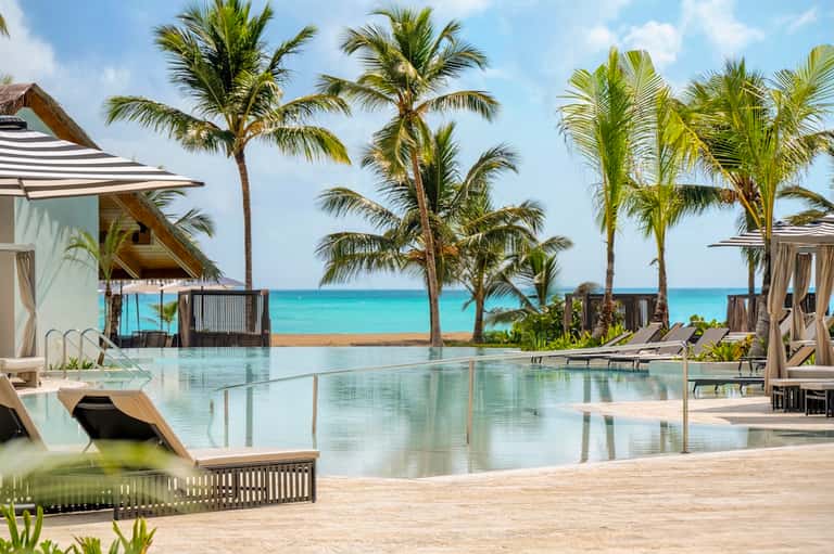 Beachfront infinity pool overlooking turquoise ocean, palm trees, cabanas with umbrellas, and sand beach