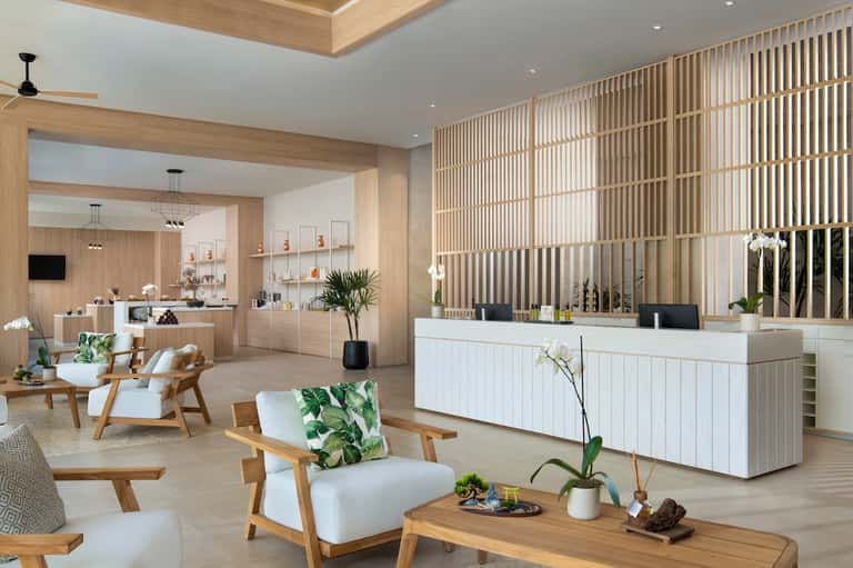 Spa reception area with light wood finishes, white reception desk, vertical slat screens, and plants
