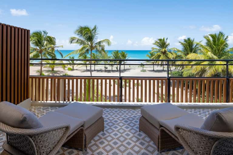 Beachfront balcony with lounge chairs overlooking palm trees, white sandy beach, and clear blue ocean water