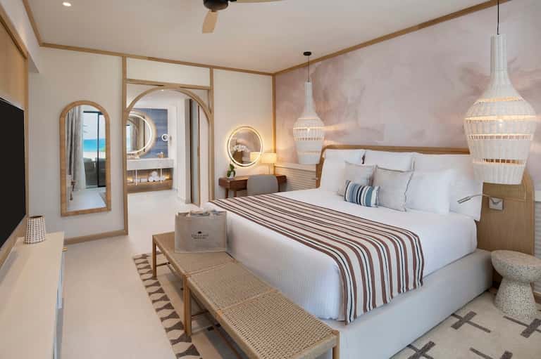 Modern beach resort bedroom with neutral tones, illuminated mirrors, pendant lights, and ocean view through open doors