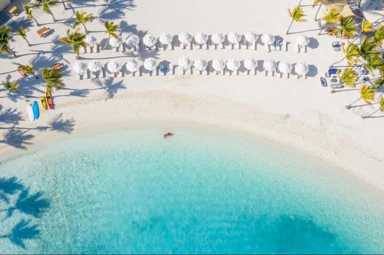 Aerial view of pristine beach with white umbrellas, lounge chairs, palm trees, and turquoise water