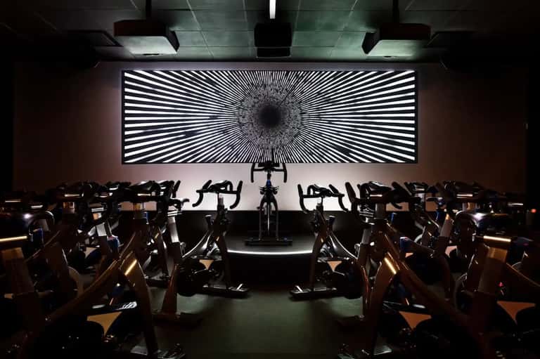 Modern fitness studio with rows of stationary bikes facing large hypnotic light display screen