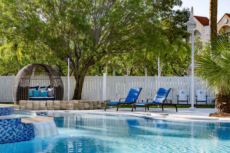 Luxury pool with waterfall feature, woven hanging chair, blue lounge chairs, white fence, and mature shade trees