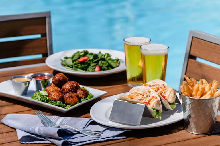 Poolside dining table with food, drinks, and ocean view; tacos, meatballs, salad, and beer