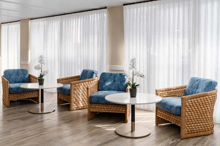 Bright lounge area with wicker chairs, blue patterned cushions, white sheer curtains, and round tables