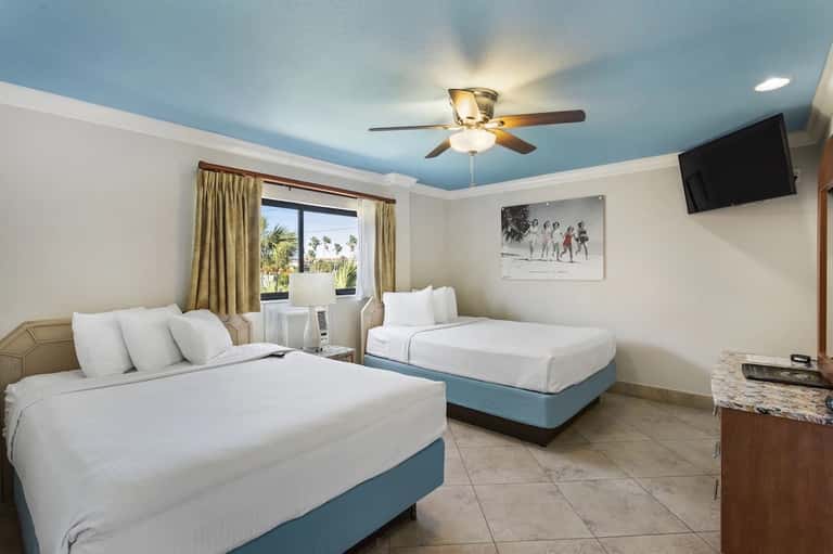 Bright hotel room with two queen beds, blue ceiling, palm tree view, and flat-screen TV