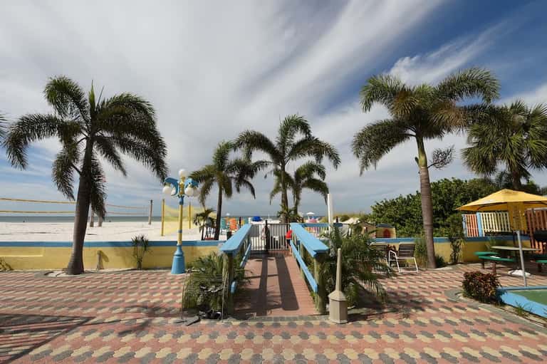 Beachfront resort grounds with palm trees, blue pathway bridge, playground, and yellow umbrellas