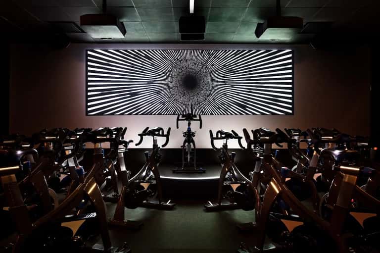 Fitness studio with rows of stationary bikes and large geometric light display wall