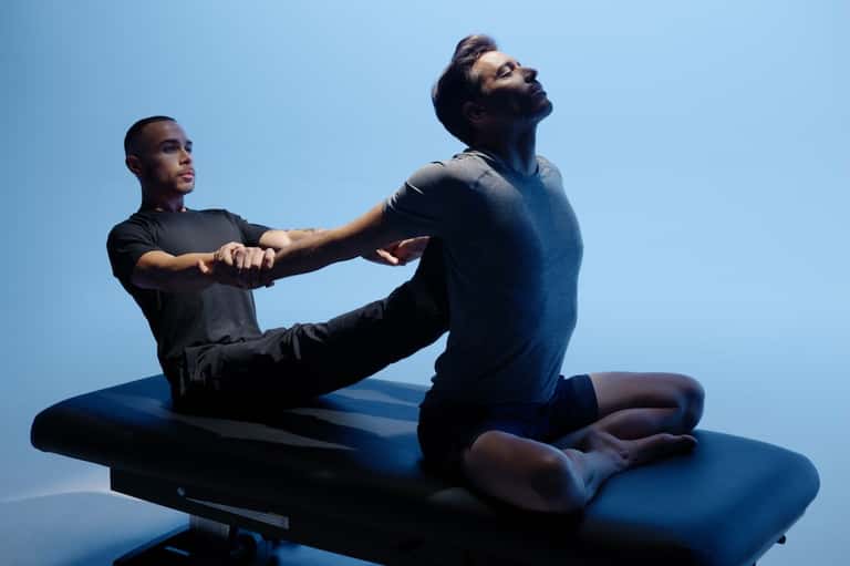 Wellness instructor assisting client in stretching pose on mat against clear blue sky background