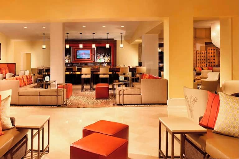 Grand hotel lobby with orange accent walls, multiple seating areas, bar, and warm ambient lighting