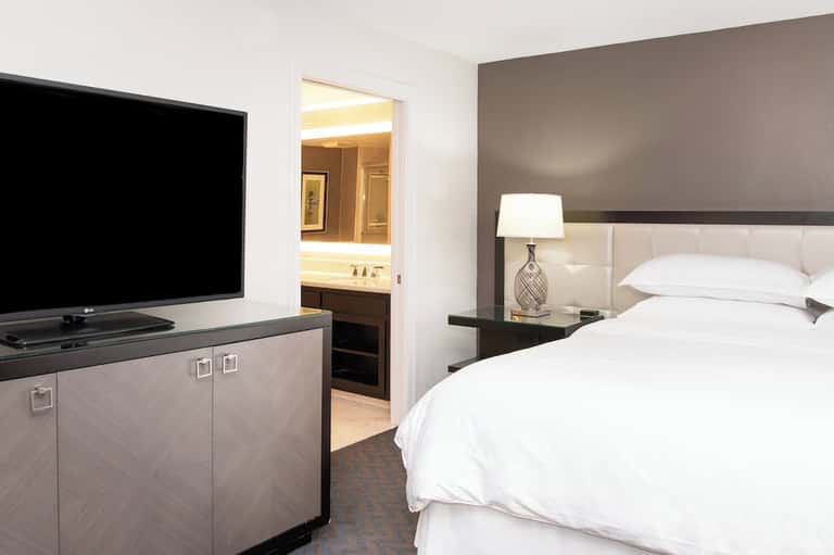 Hotel bedroom with TV, white bedding, gray headboard, and open bathroom door