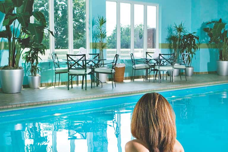 Indoor swimming pool with turquoise water, large windows overlooking greenery, and seating areas with tropical plants