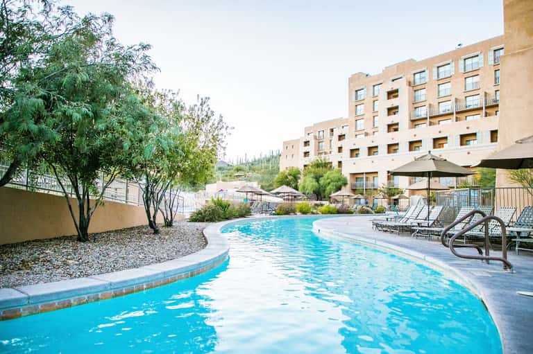 Resort courtyard with curved swimming pool, multi-story tan building, lounge chairs, and landscaping