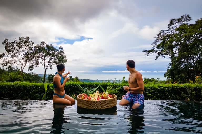 Two people in infinity pool sharing floating fruit basket with lush tropical landscape background