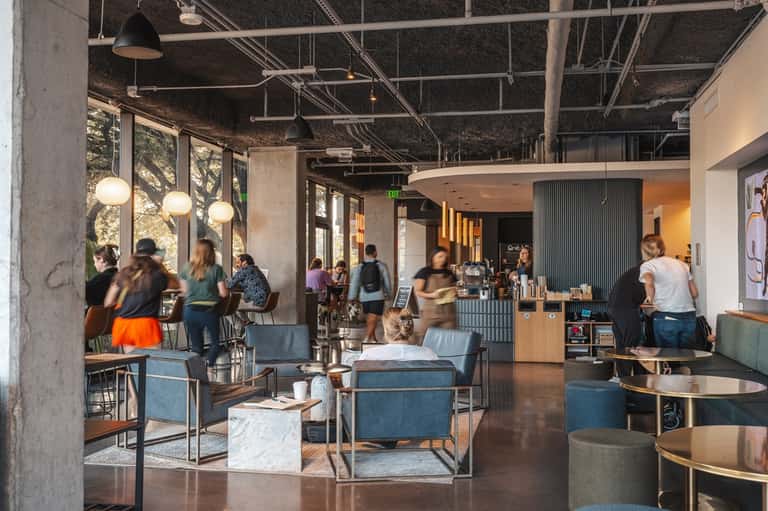 Modern hotel lobby with exposed ceiling, pendant lighting, concrete walls, and guests at bar counter