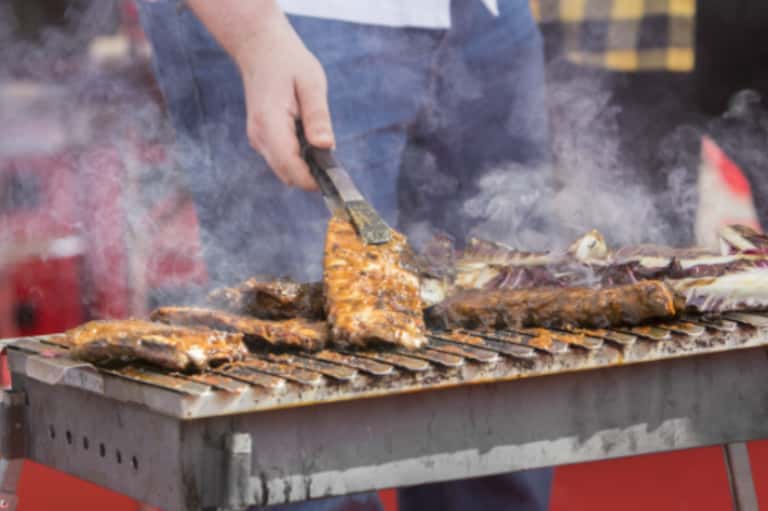 Person grilling fish over smoky outdoor fire pit with hands and cooking utensils visible