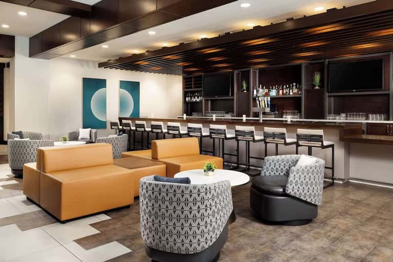 Contemporary hotel lounge with orange sofa, patterned accent chairs, full bar, and modern lighting fixtures